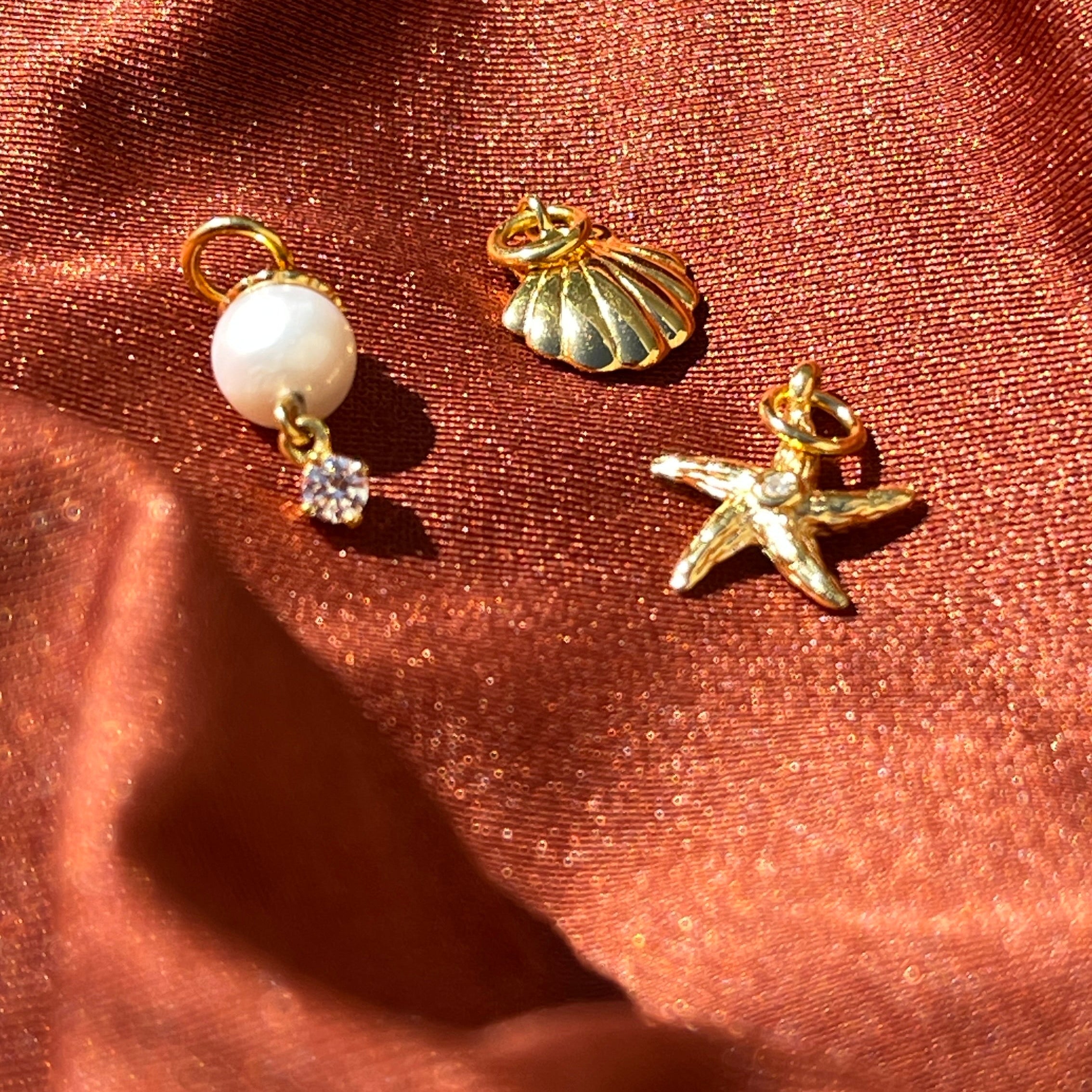 Zaira Jewels ocean-themed 18k gold vermeil charms, including a white pearl, a golden seashell, and a starfish, displayed on luxurious terracotta silk fabric.