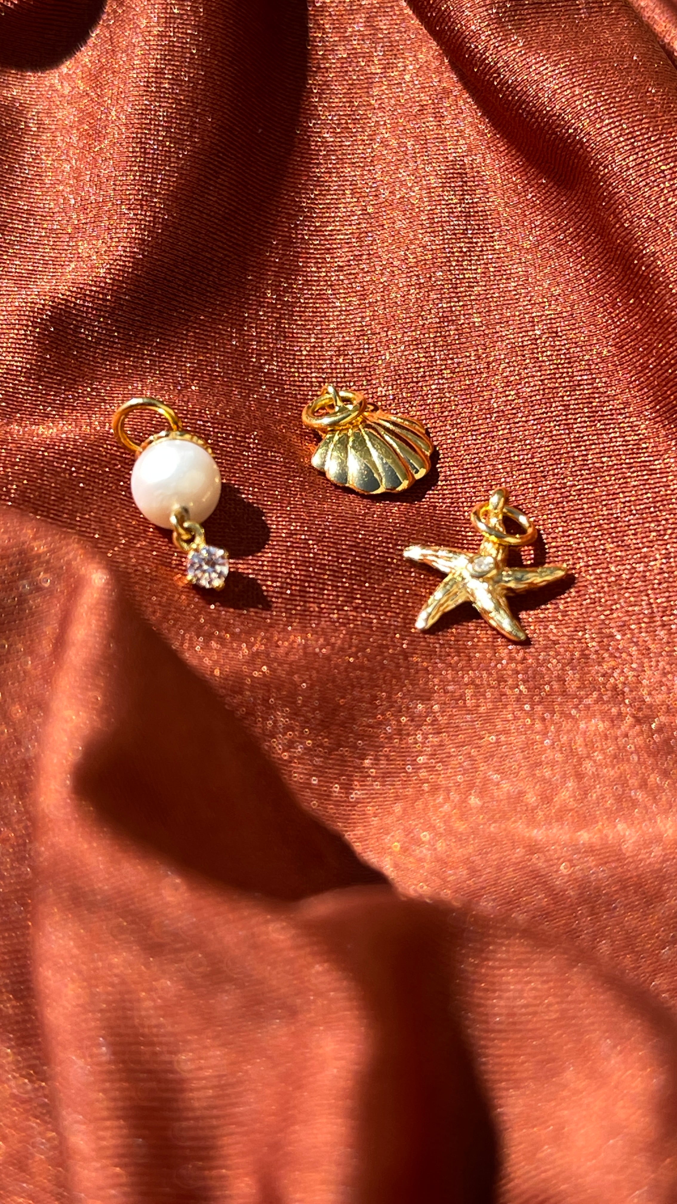 Zaira Jewels ocean-themed 18k gold vermeil charms, including a white pearl, a golden seashell, and a starfish, displayed on luxurious terracotta silk fabric.